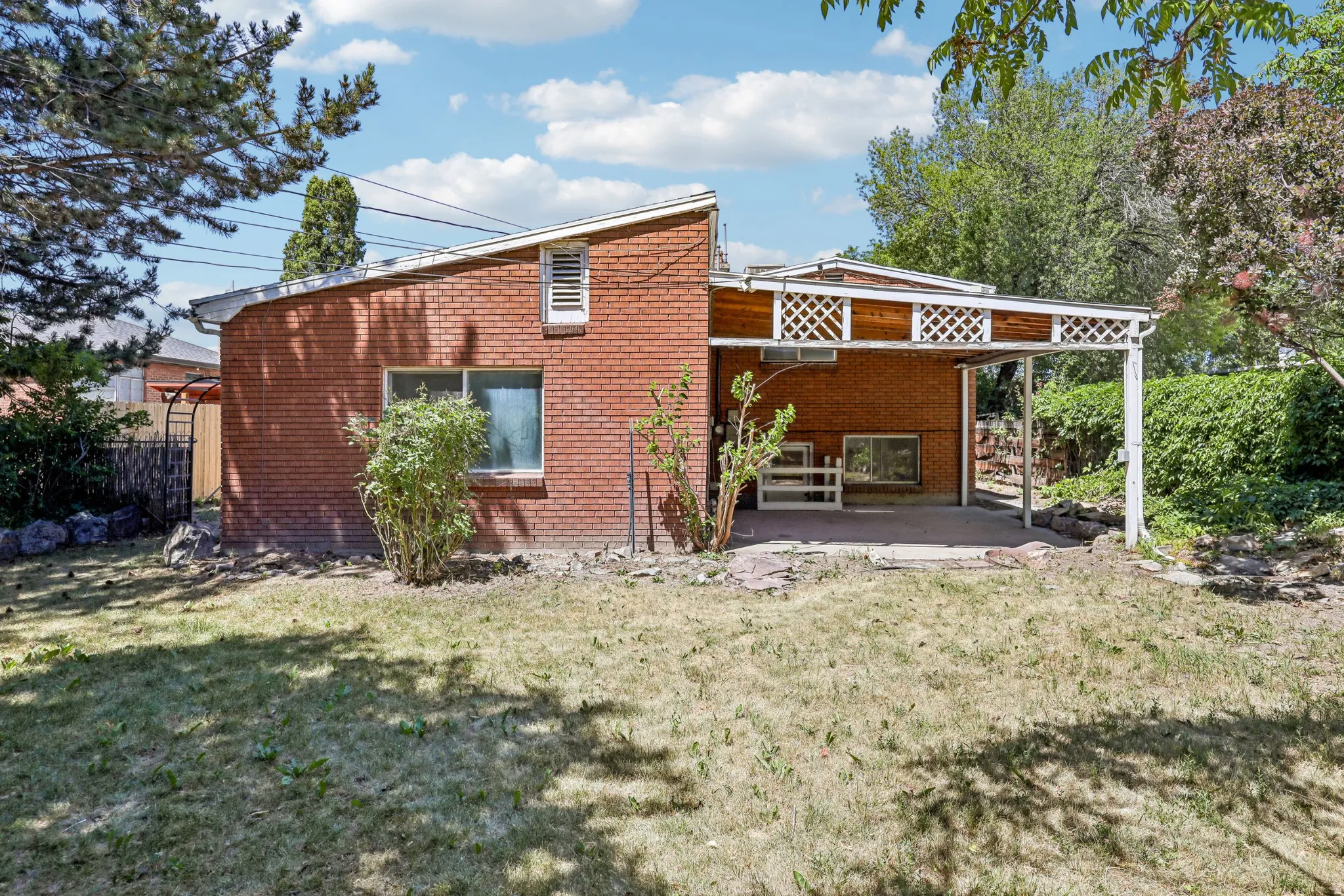 Brick exterior featuring an integrated covered patio with lattice detailing, a large window, and mature perimeter foliage