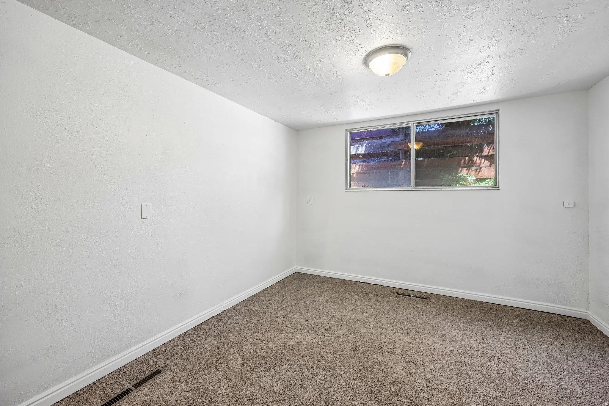 Neutral-toned carpeted room featuring a window, white walls, and a ceiling-mounted light fixture