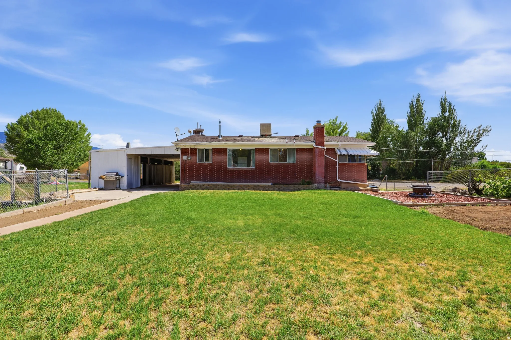 Brick exterior residence featuring a carport, green lawn, gravel patio, and a chain-link fenced area
