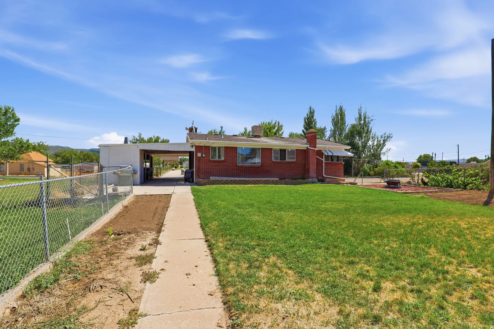 Brick exterior residence featuring a green lawn, concrete walkway, chain-link fencing, and an attached carport
