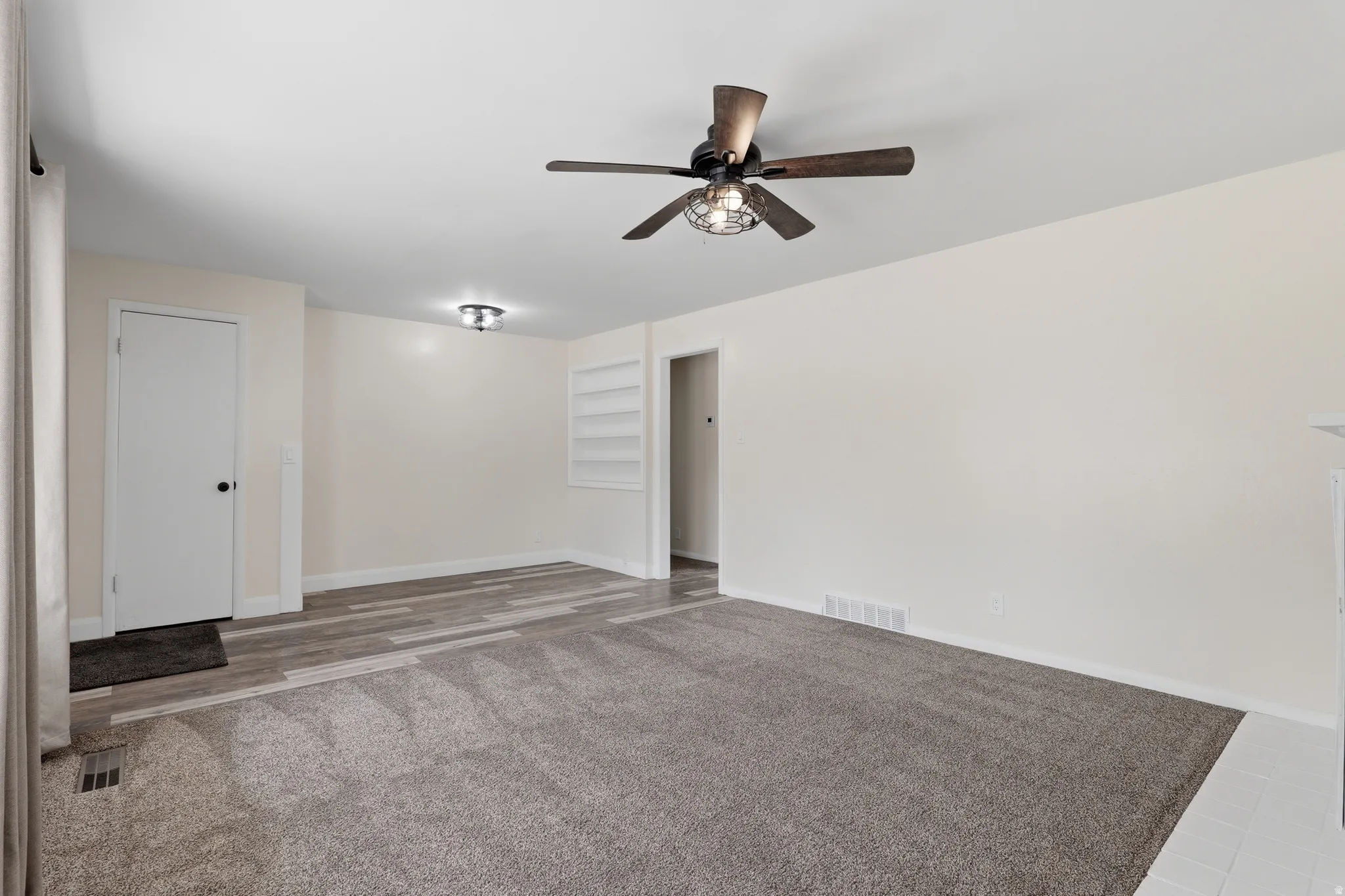 Spacious room featuring neutral wall tones, a modern ceiling fan with integrated lighting, a recessed built-in shelving unit, and a combination of carpet and wood-finish flooring