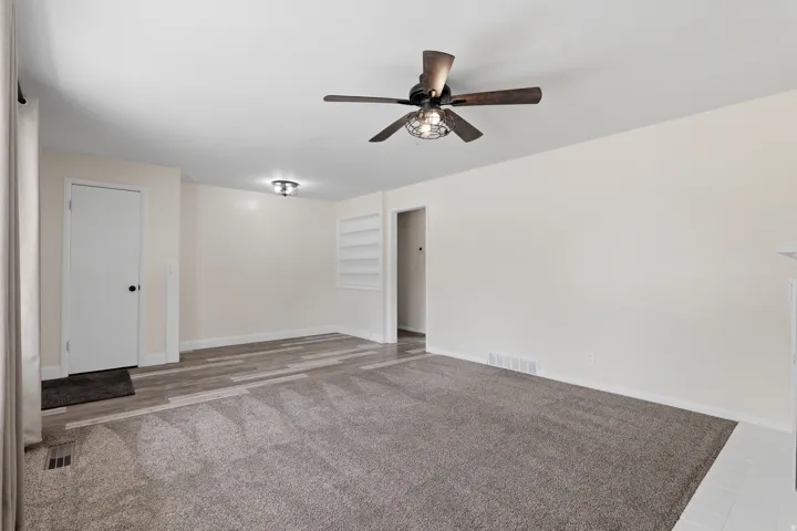 Spacious room featuring neutral wall tones, a modern ceiling fan with integrated lighting, a recessed built-in shelving unit, and a combination of carpet and wood-finish flooring
