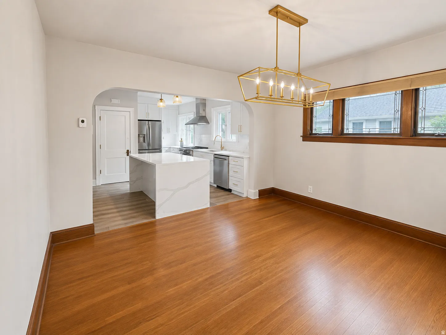 Hardwood flooring, arched doorway, kitchen island with waterfall countertop, stainless steel appliances, and a linear chandelier