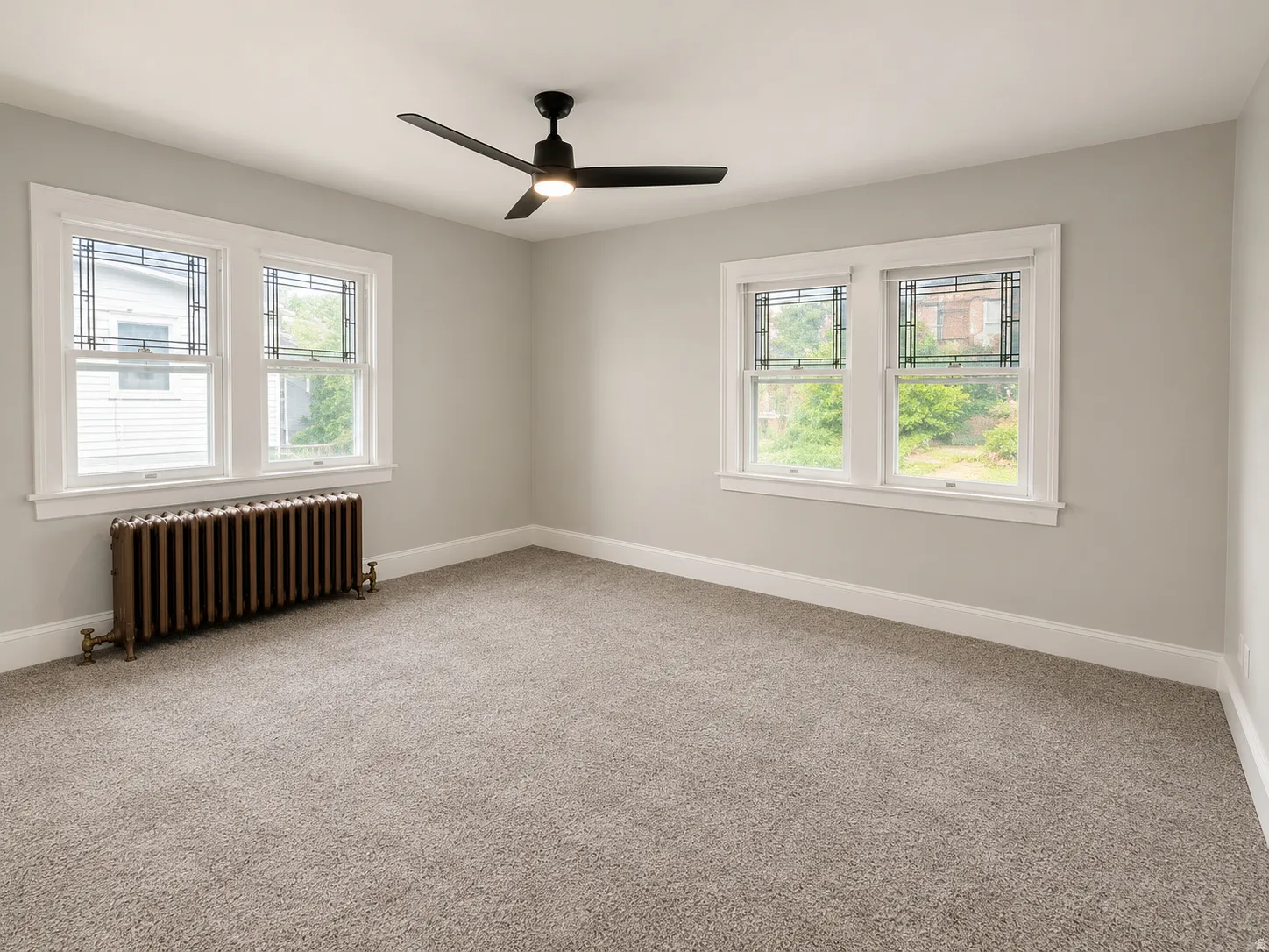 Light gray wall paint, gray carpet flooring, matte black ceiling fan, white window trim, and a cast iron radiator