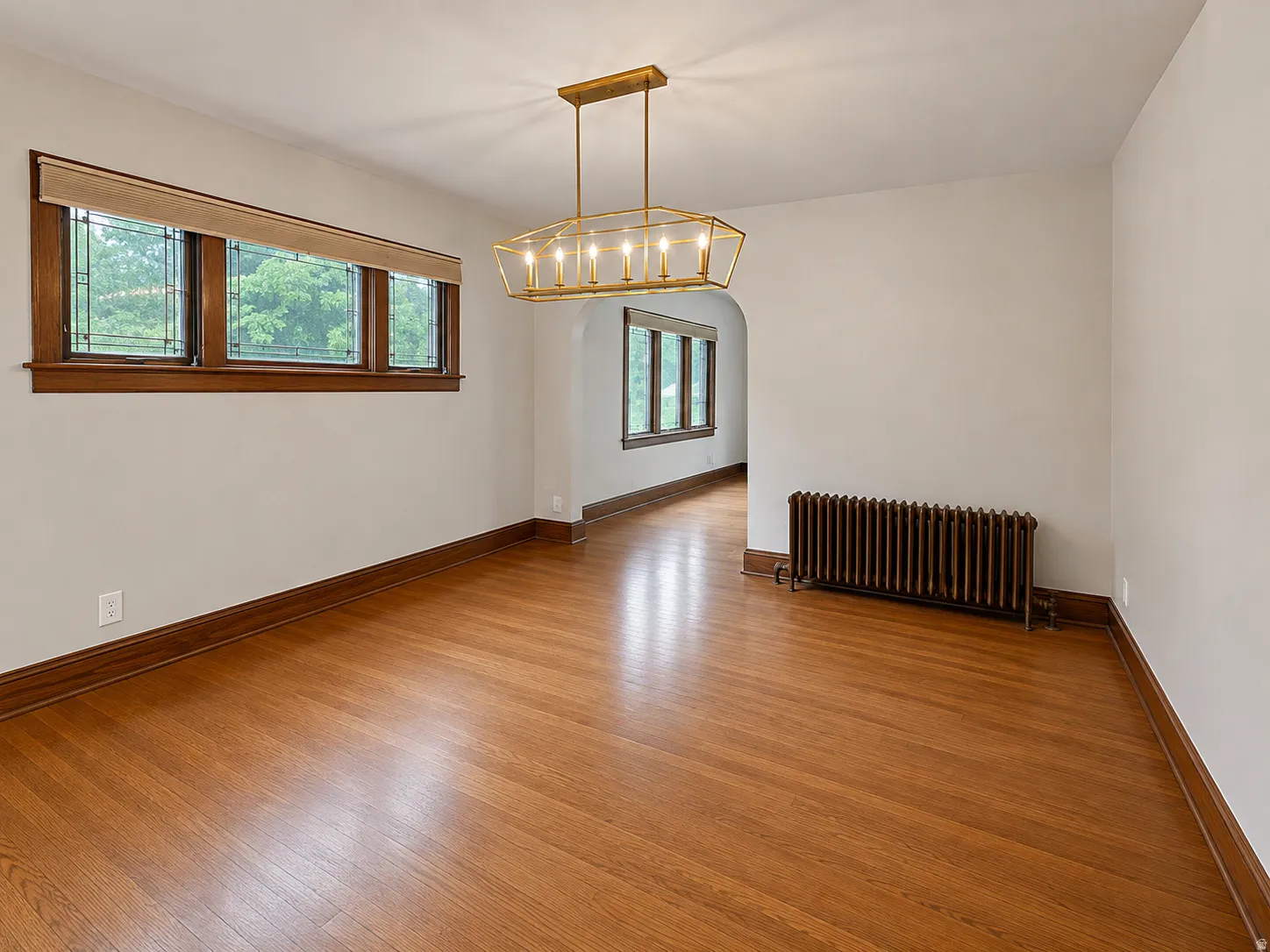 Hardwood flooring, decorative chandelier, wood-trimmed windows with privacy film, traditional radiator, and wood baseboards