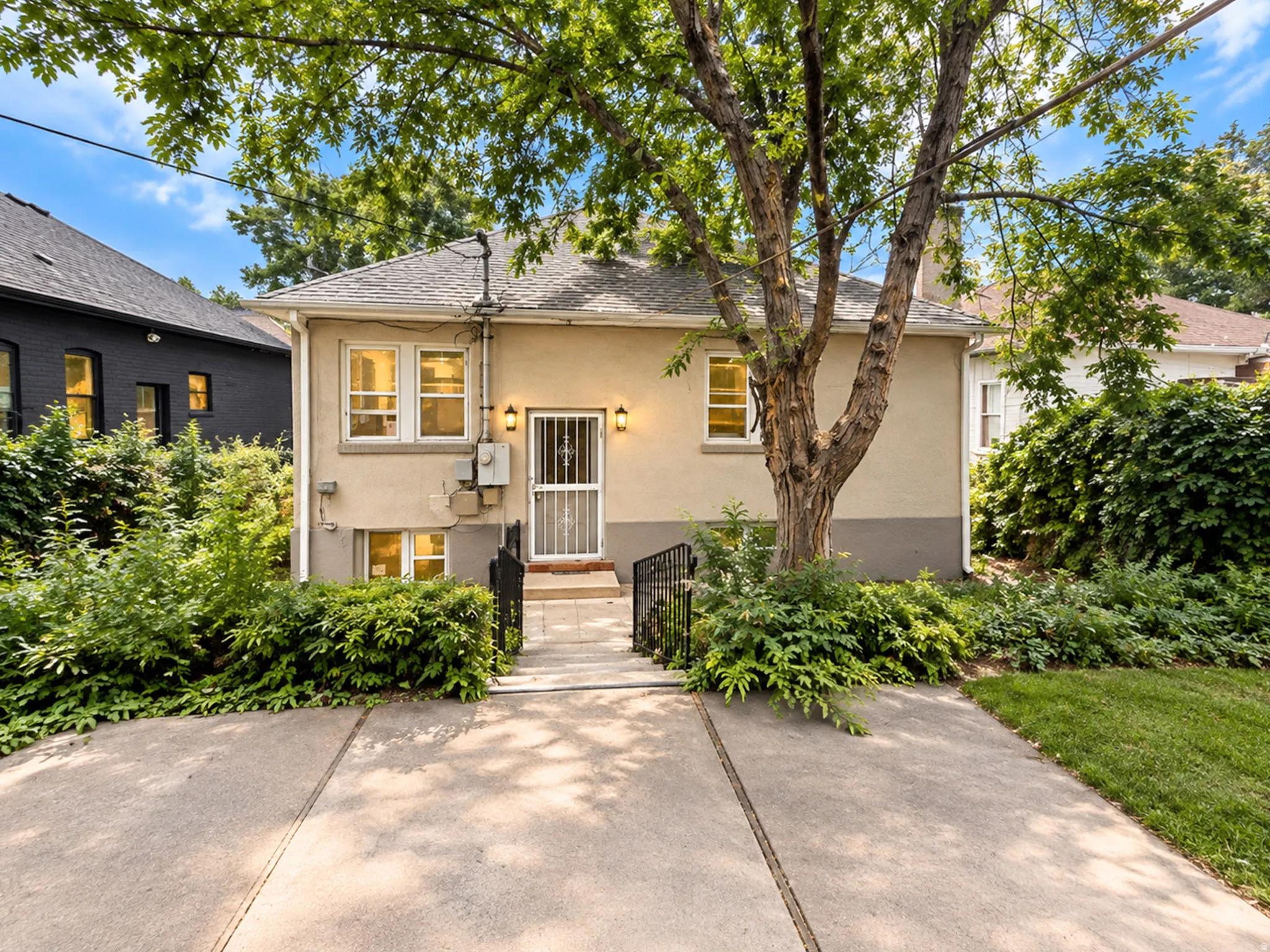 Stucco exterior with a pitched roof, concrete walkway, wrought iron railings, and mature landscaping