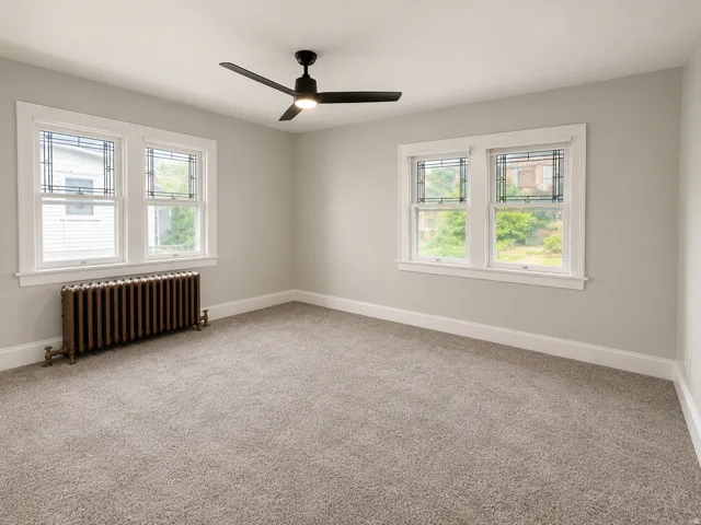 Light gray wall paint, gray carpet flooring, matte black ceiling fan, white window trim, and a cast iron radiator