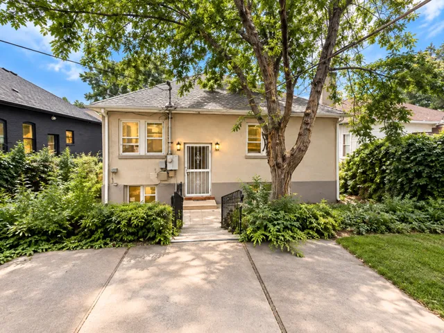 Stucco exterior with a pitched roof, concrete walkway, wrought iron railings, and mature landscaping