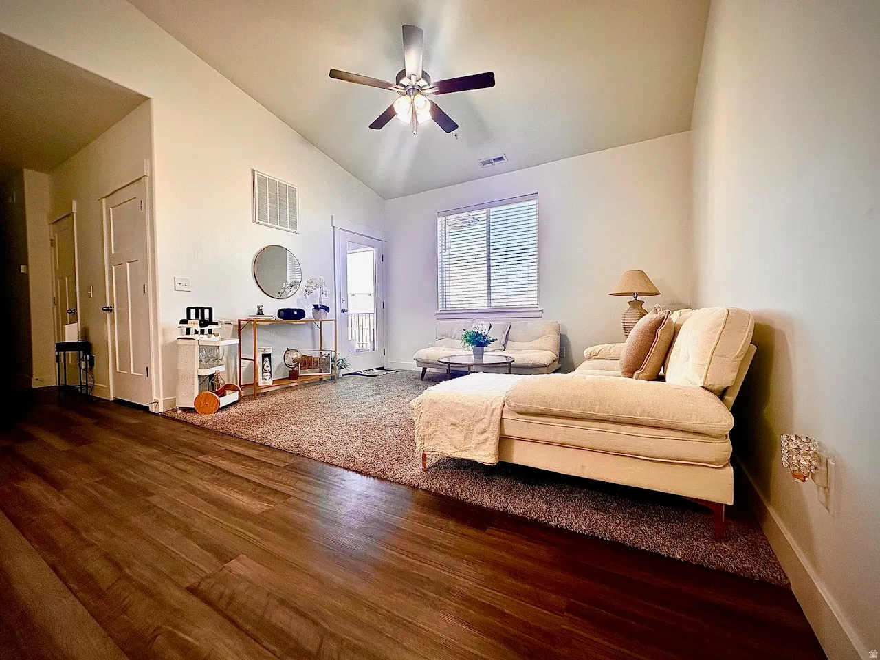 Vaulted ceiling living area featuring wood-finish flooring, a ceiling fan with integrated lighting, and a large window with blinds
