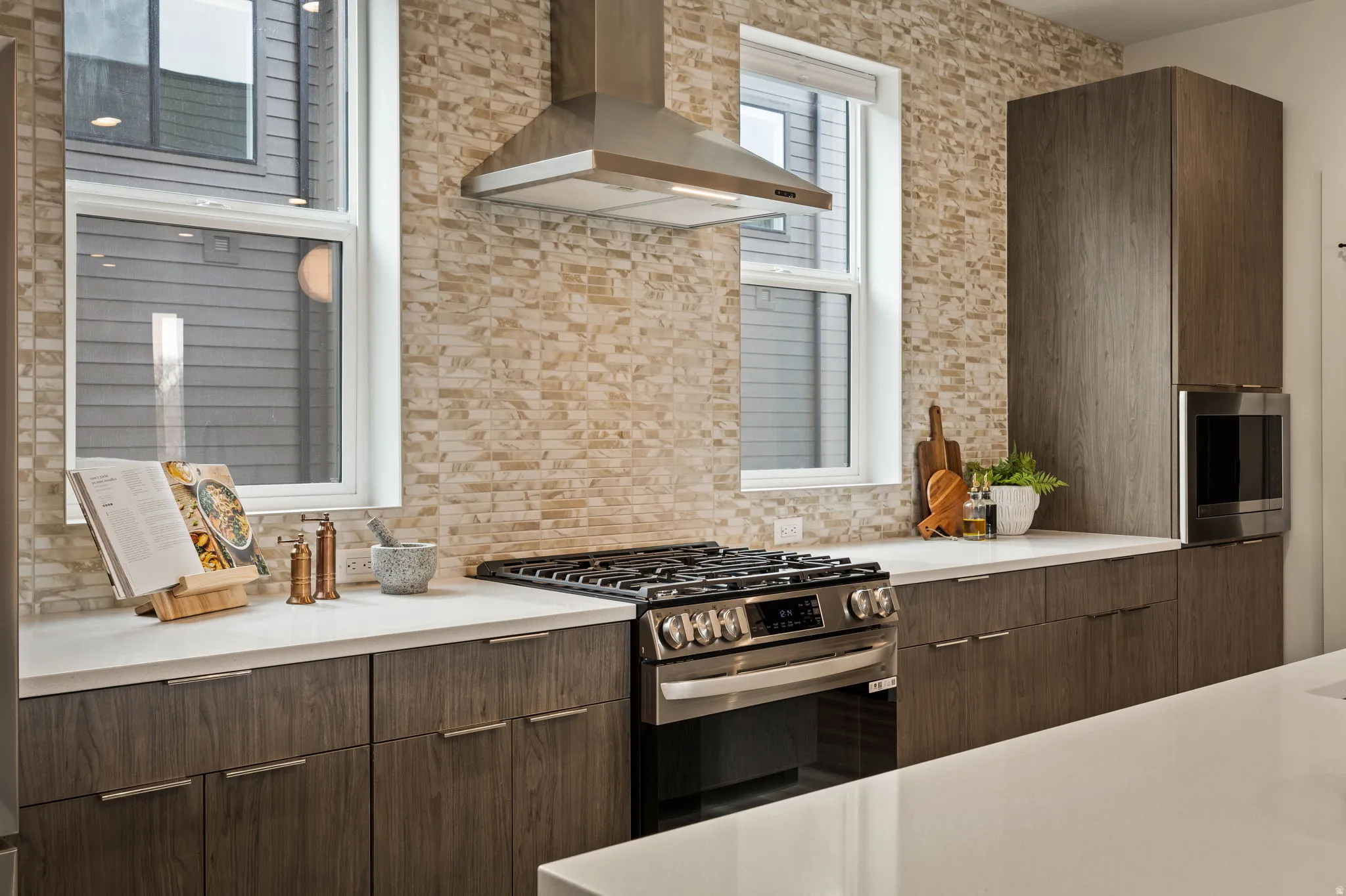 Modern kitchen featuring a stainless steel range with a coordinating hood, stone-finish backsplash, wood-grain cabinetry, light-toned countertops, and dual windows