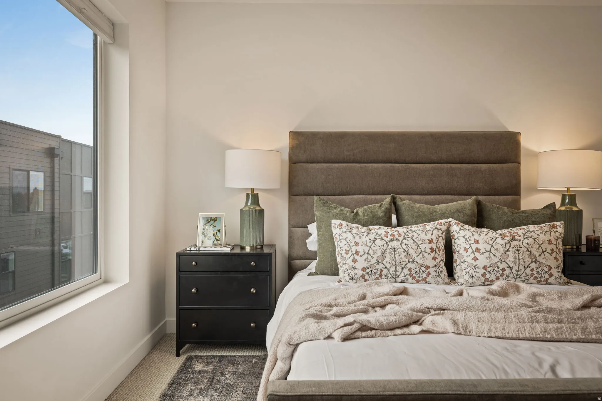 Bedroom featuring a large window, a paneled upholstered headboard, a dark wood nightstand with drawer pulls, and wall-to-wall carpeting