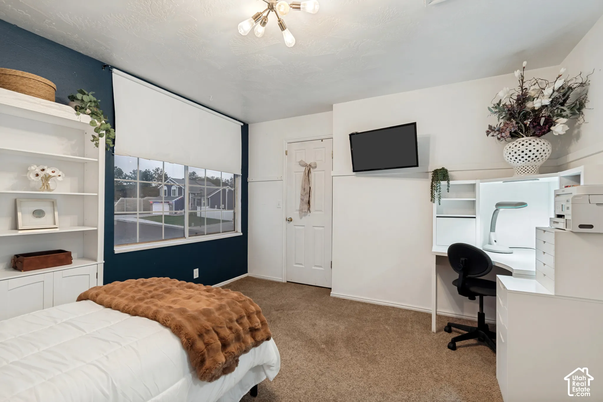 Room featuring a multi-pane window with roller shade, built-in shelving, a recessed desk with integrated lighting, and neutral carpeting