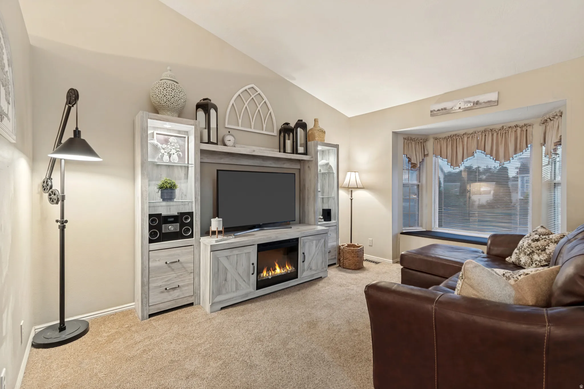 Living area featuring a vaulted ceiling, light beige carpeting, a built-in media console with an electric fireplace, and a large bay window with an integrated bench seat