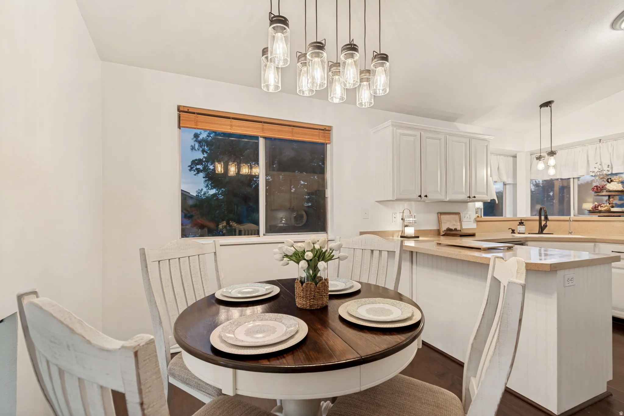 Bright dining area featuring a modern chandelier with multiple exposed bulbs, a large window with wood blinds, white shaker-style cabinetry, light wood-finish countertops, and dark wood-finish flooring