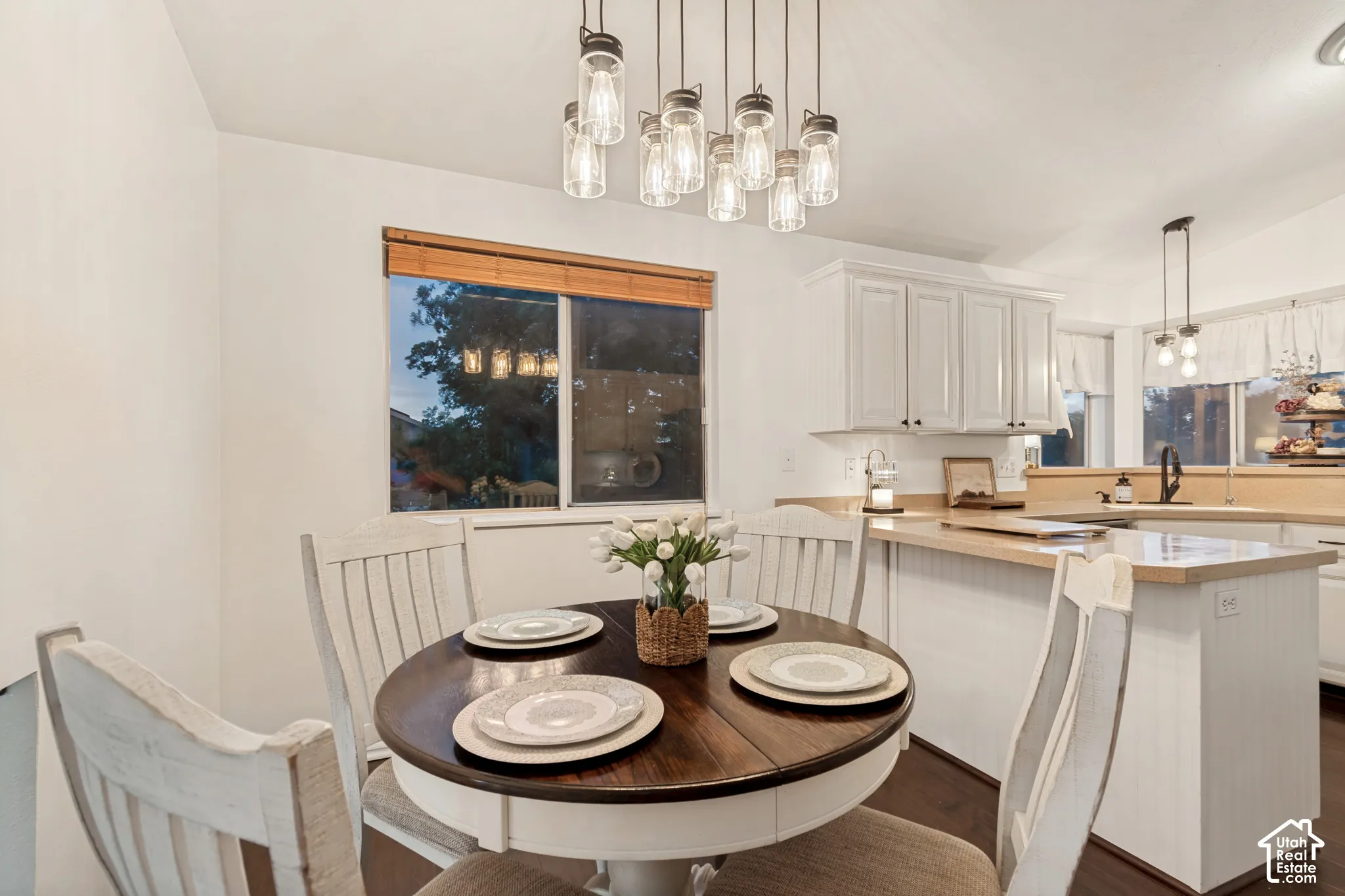 Bright dining area featuring a modern chandelier with multiple exposed bulbs, a large window with wood blinds, white shaker-style cabinetry, light wood-finish countertops, and dark wood-finish flooring