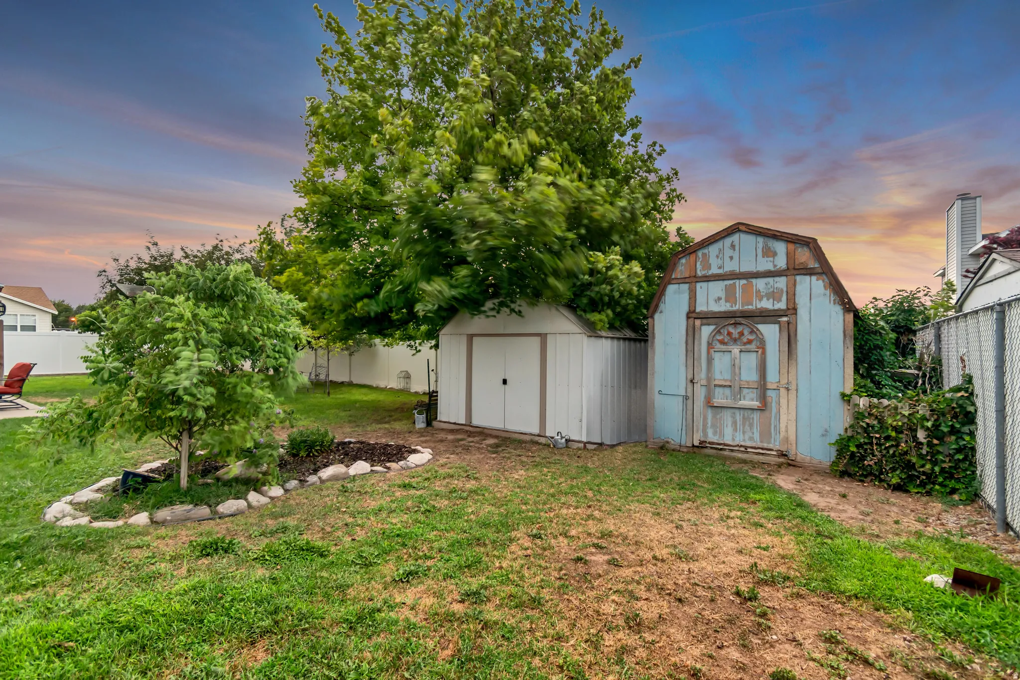 Expansive backyard featuring two sheds, mature trees, a white privacy fence, and a decorative rock-lined planting bed