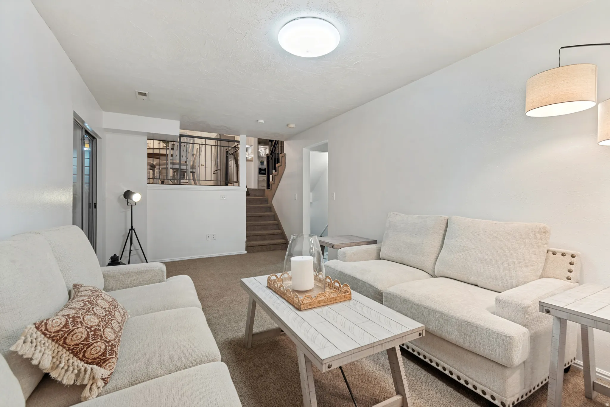 Carpeted living area featuring white walls, a flush-mount ceiling light, and a staircase with a black metal railing