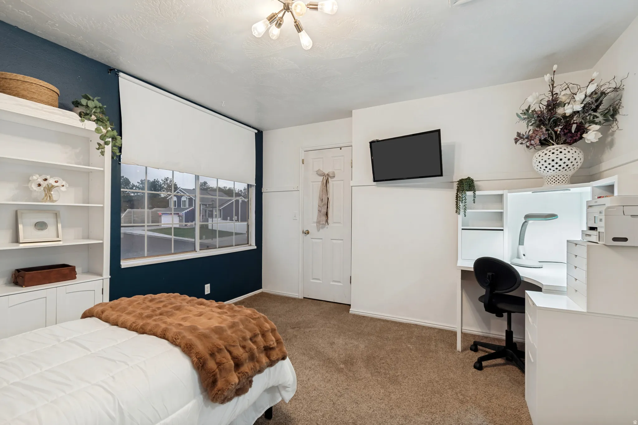 Room featuring a multi-pane window with roller shade, built-in shelving, a recessed desk with integrated lighting, and neutral carpeting