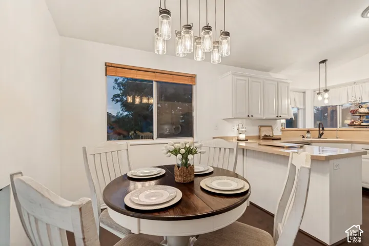 Bright dining area featuring a modern chandelier with multiple exposed bulbs, a large window with wood blinds, white shaker-style cabinetry, light wood-finish countertops, and dark wood-finish flooring