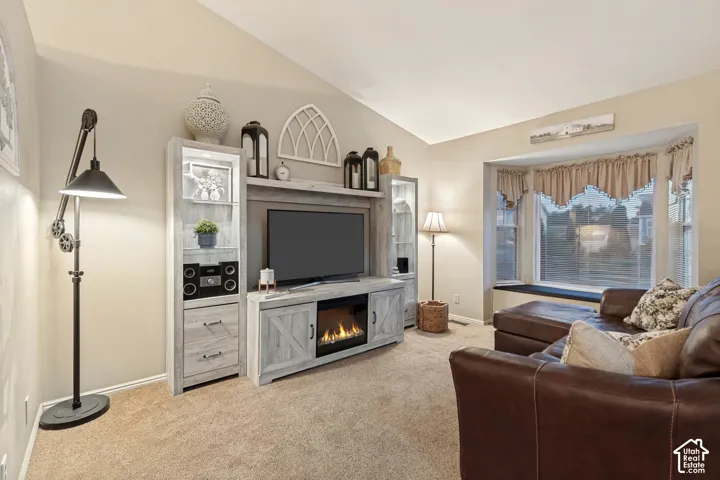 Living area featuring a vaulted ceiling, light beige carpeting, a built-in media console with an electric fireplace, and a large bay window with an integrated bench seat