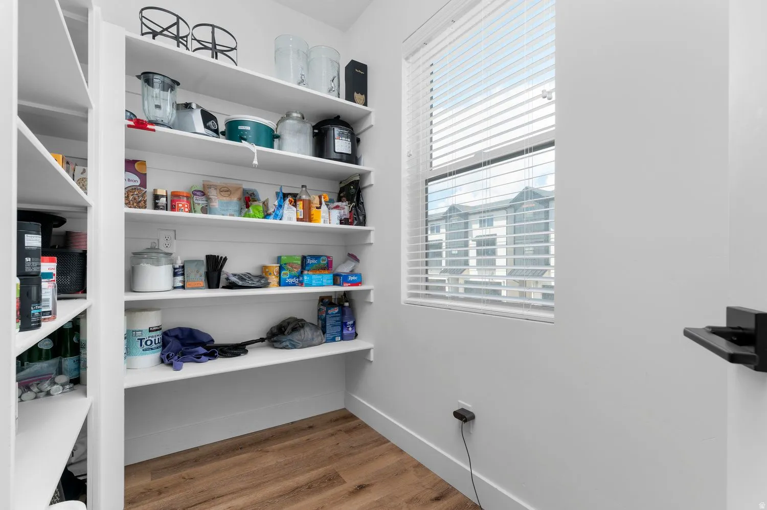 Walk-in pantry featuring extensive built-in shelving, wood-finish flooring, a window with blinds, and white painted walls