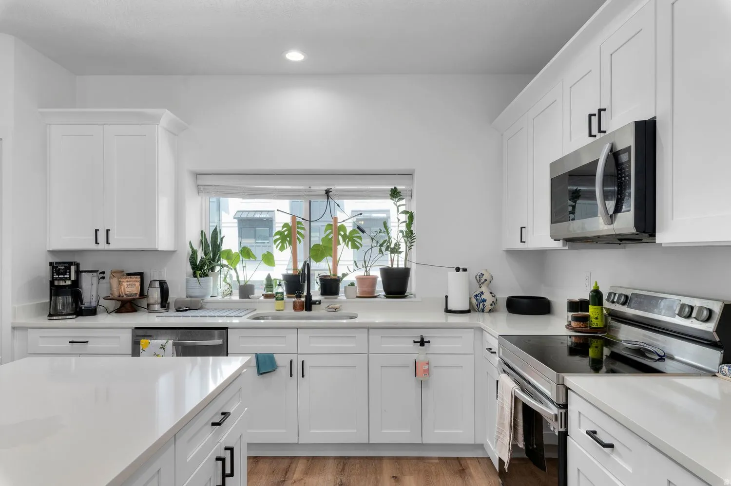 Bright kitchen featuring white shaker cabinetry, stainless steel appliances, light countertops, wood-finish flooring, and a large window above the sink