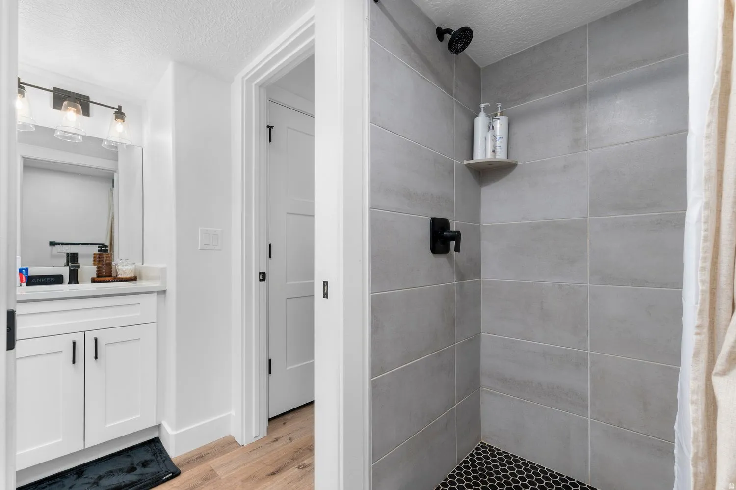 Contemporary bathroom featuring a tiled shower with black hexagonal floor tiles and matte black fixtures, a floating corner shelf, white vanity with white countertop, and wood-finish flooring