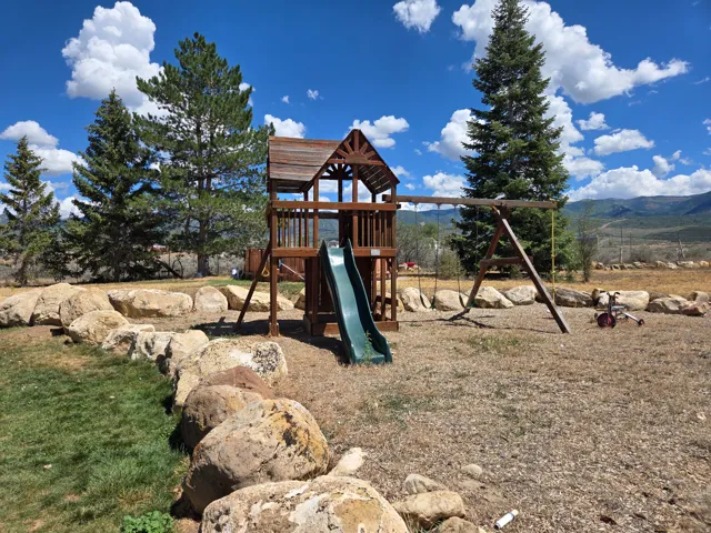 Expansive backyard featuring a wood-toned play structure with a slide and swing set, mature evergreen trees, and a natural rock retaining wall