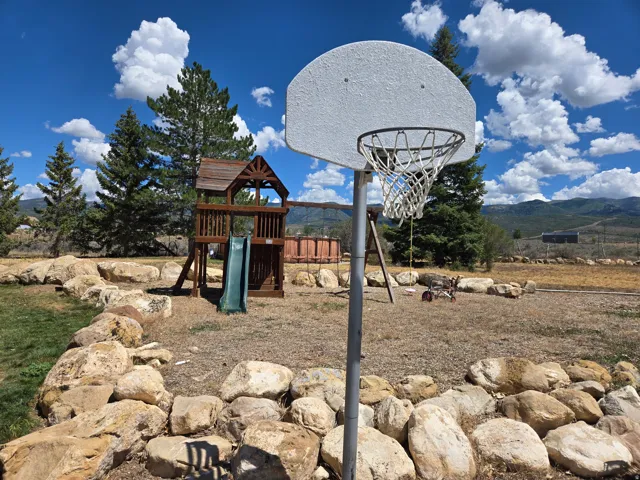 Outdoor recreational area featuring a basketball hoop, wooden play structure with slide, and natural rock landscaping