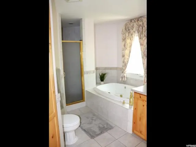 Bathroom featuring a built-in jetted tub, a frosted glass shower enclosure with gold-tone framing, a window with decorative valance and drapes, light-toned tile flooring, and a toilet