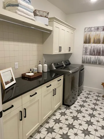 Utility room featuring patterned floor tiling, cream shaker-style cabinetry with matte black hardware, black stone countertop, a full-height tile backsplash, and an overhead shelf with integrated lighting