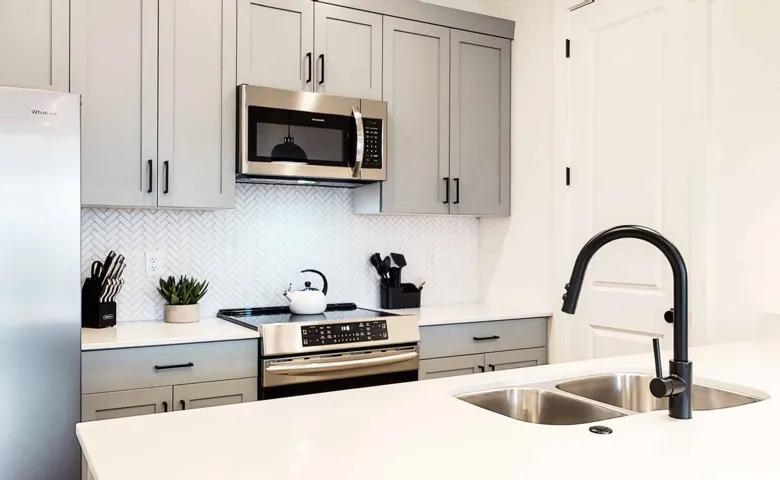 Modern kitchen featuring light gray cabinetry with black hardware, stainless steel appliances, white herringbone tile backsplash, and light-toned countertops