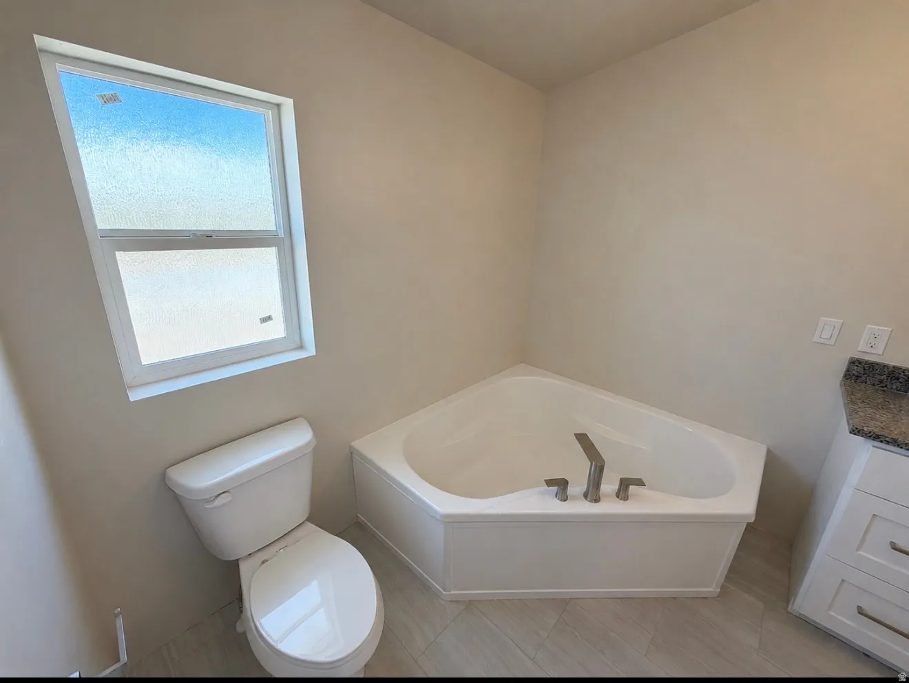 Corner soaking tub with brushed nickel fixtures, accompanied by a white toilet and a single-hung window with privacy glass