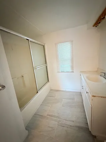 Bathroom featuring a white bathtub with sliding glass enclosure, a single vanity with a white countertop, and light-toned tile flooring
