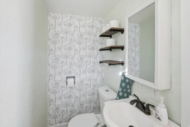 Powder room featuring a floral accent wall, wall-mounted wood-finish shelving, a white vanity with an integrated sink, a dark bronze-finish faucet, and a mirrored medicine cabinet