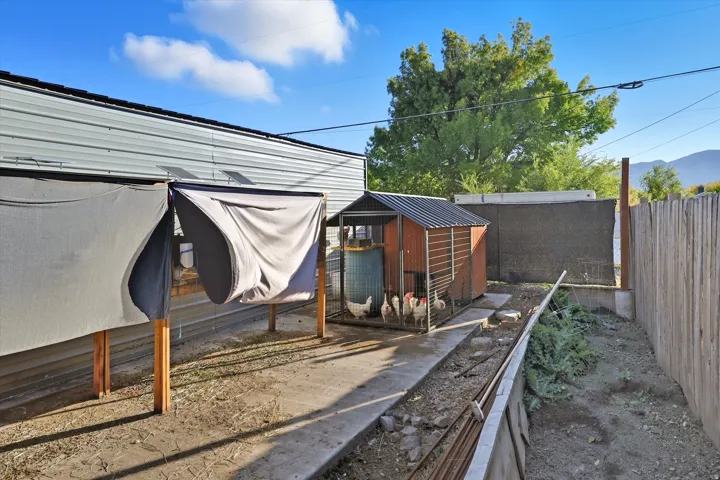Outdoor space featuring a chicken coop with a metal roof, a concrete pathway, a wood fence, and mature trees in the background