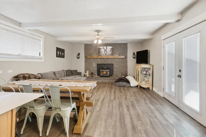 Expansive living area featuring a stone-clad fireplace with a wood-burning stove, wood-finish flooring, exposed ceiling beams, and dual French doors with integrated blinds
