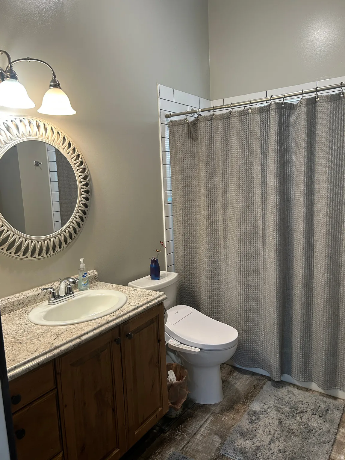 Bathroom featuring a wood-finish vanity with an integrated sink and light-toned countertop, a decorative round mirror, and a shower area with white subway tile surround