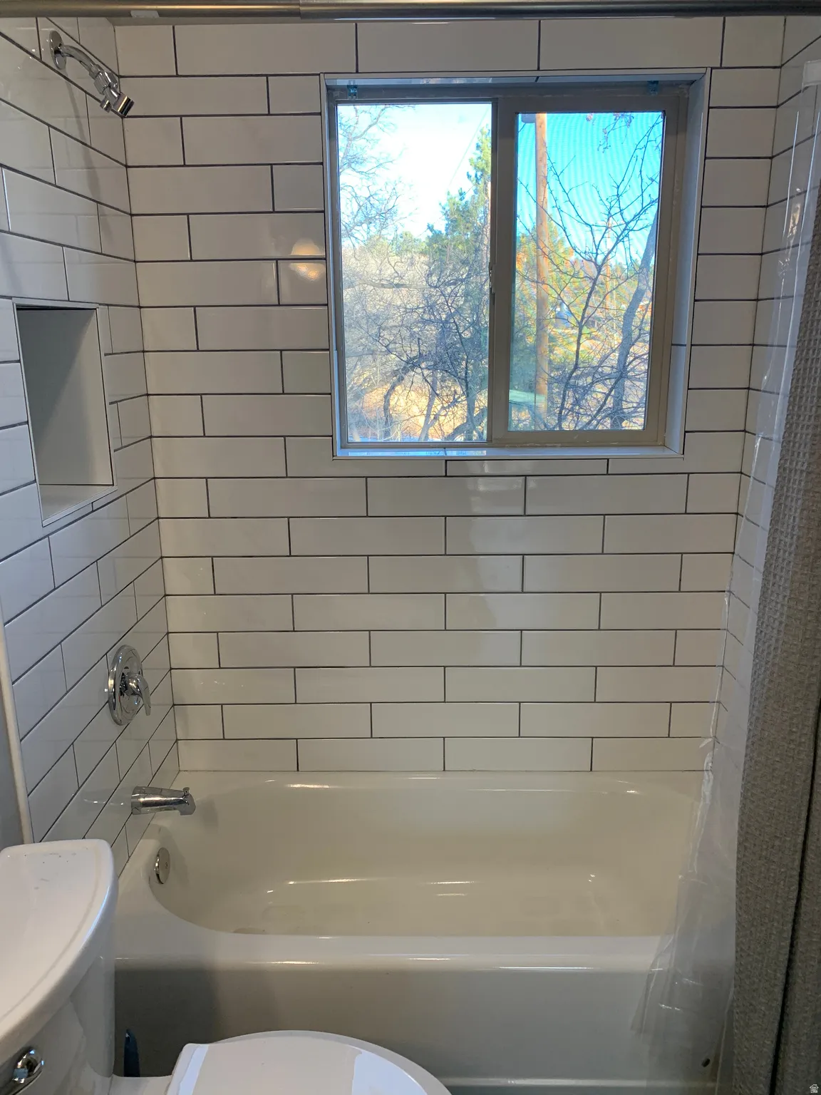 Bathtub with subway tile surround, featuring a built-in shower niche, chrome fixtures, and a window