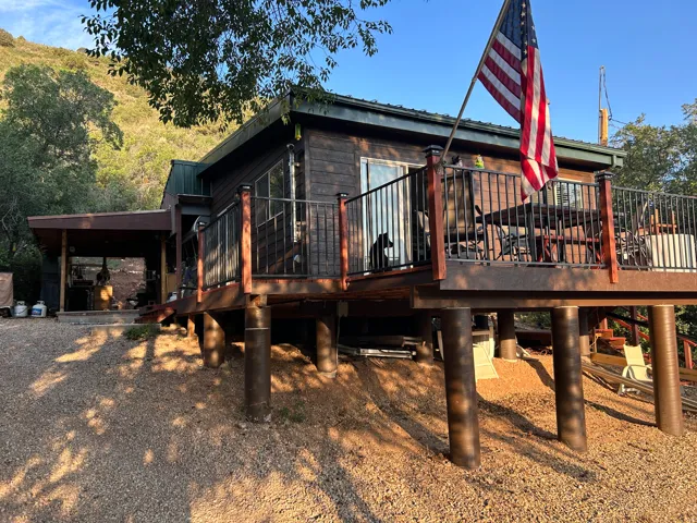 Elevated wood-sided home featuring a spacious outdoor deck with metal railings, a covered patio area, and a gravel ground surface