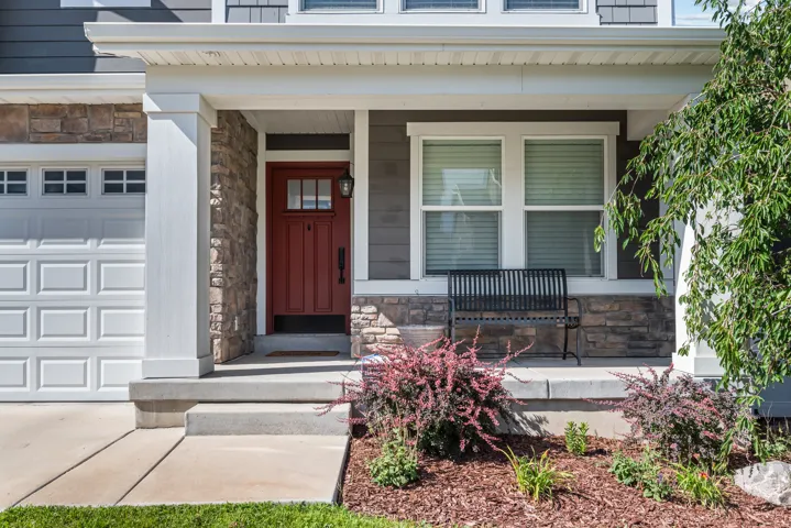 Red entry door with transom window and exterior sconce lighting, stone veneer accents, white panel garage door, concrete porch, and manicured landscaping