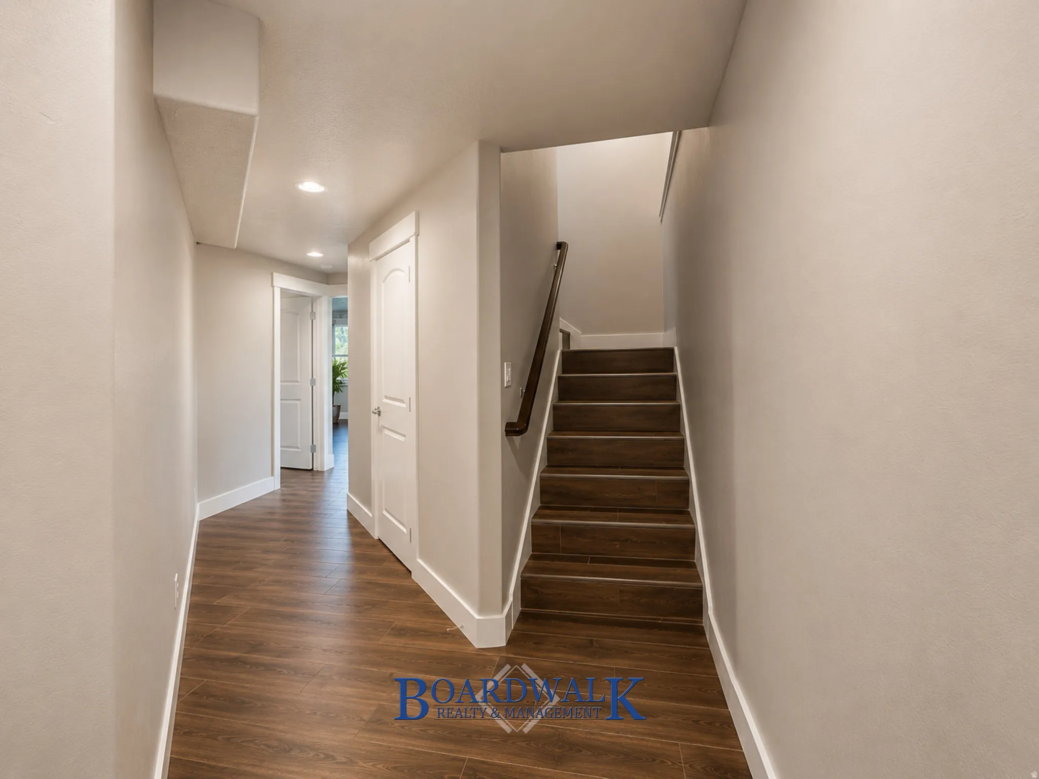 Hallway with recessed lighting, wood-finish flooring, white trim, and a staircase with a dark wood handrail