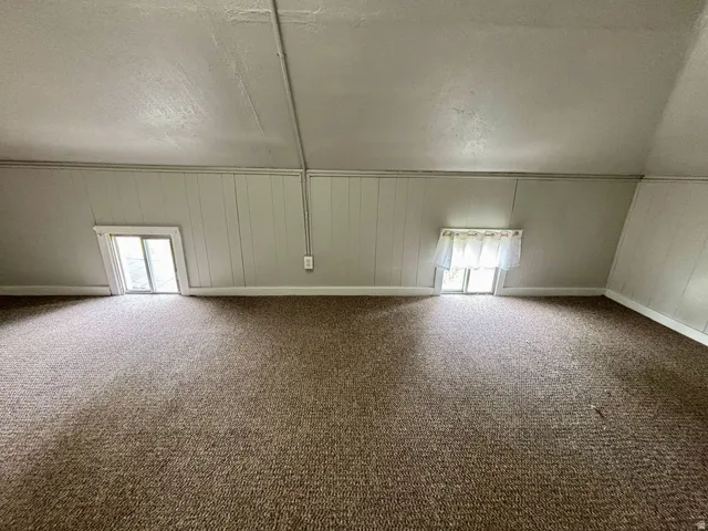 Carpeted attic space featuring sloped ceilings, two windows, and vertical paneling on perimeter walls