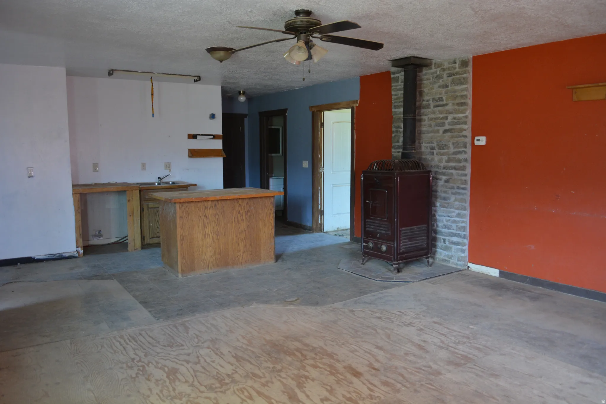 Living area featuring a floor-mounted wood stove with a stone surround, a ceiling fan, and exposed subflooring