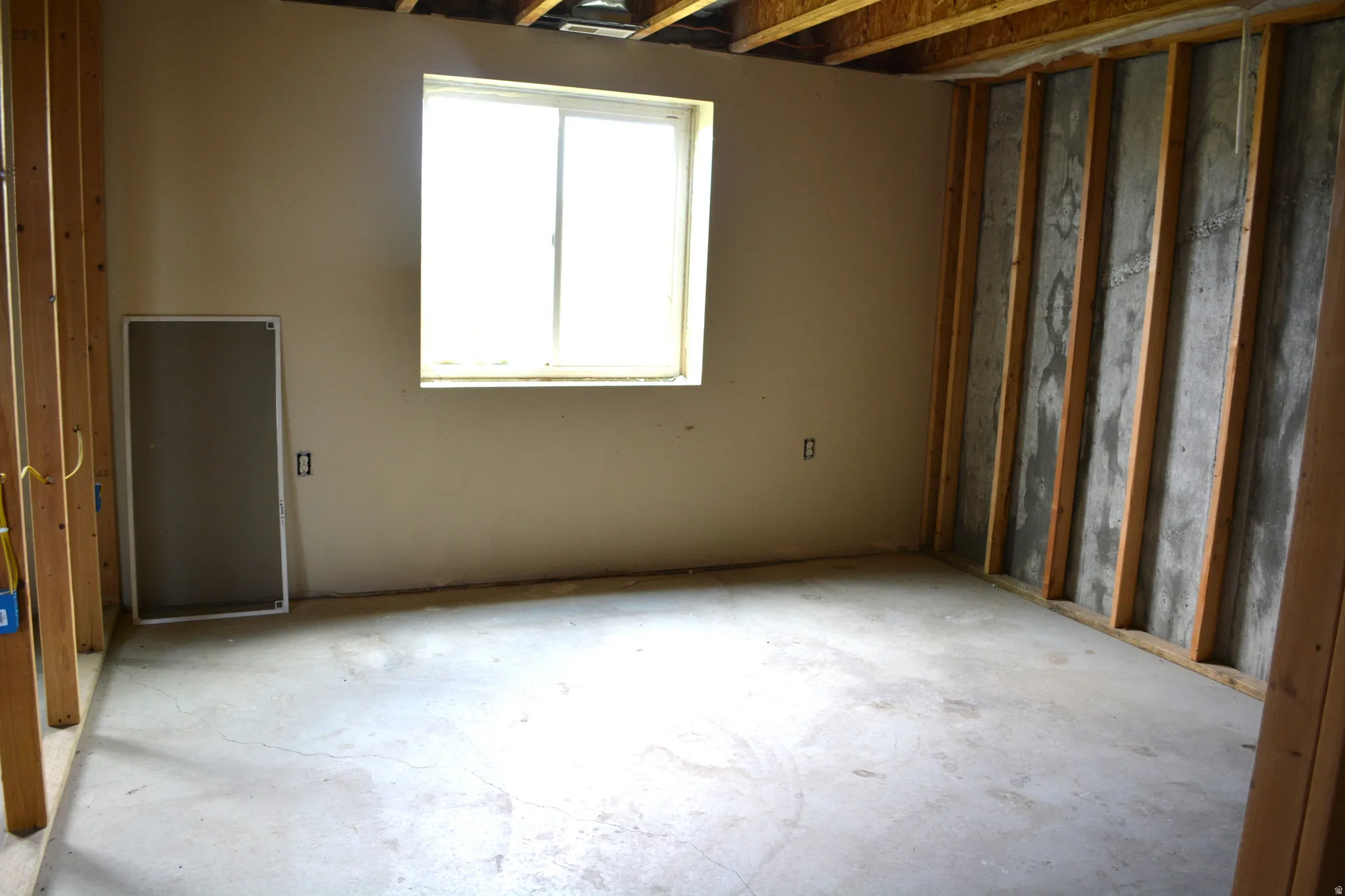 Unfinished basement room featuring a window, concrete flooring, wooden wall studs, and exposed ceiling joists