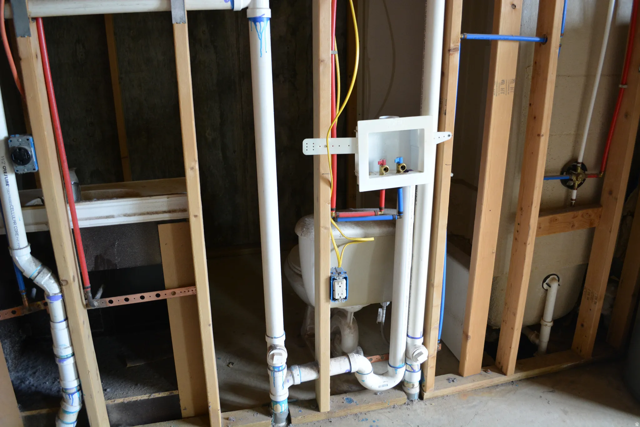Utility area framing with exposed plumbing lines for water supply and drainage, electrical wiring, and a recessed washing machine hookup box