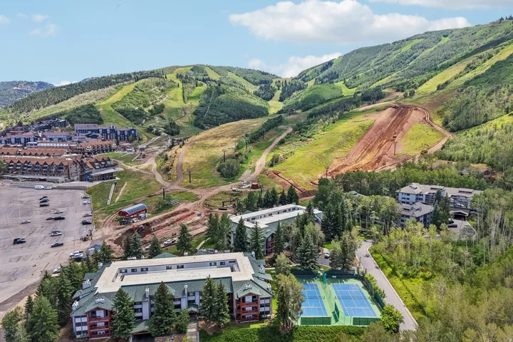 Expansive mountain setting featuring buildings with pitched roofs, surrounded by dense trees, and a tennis court