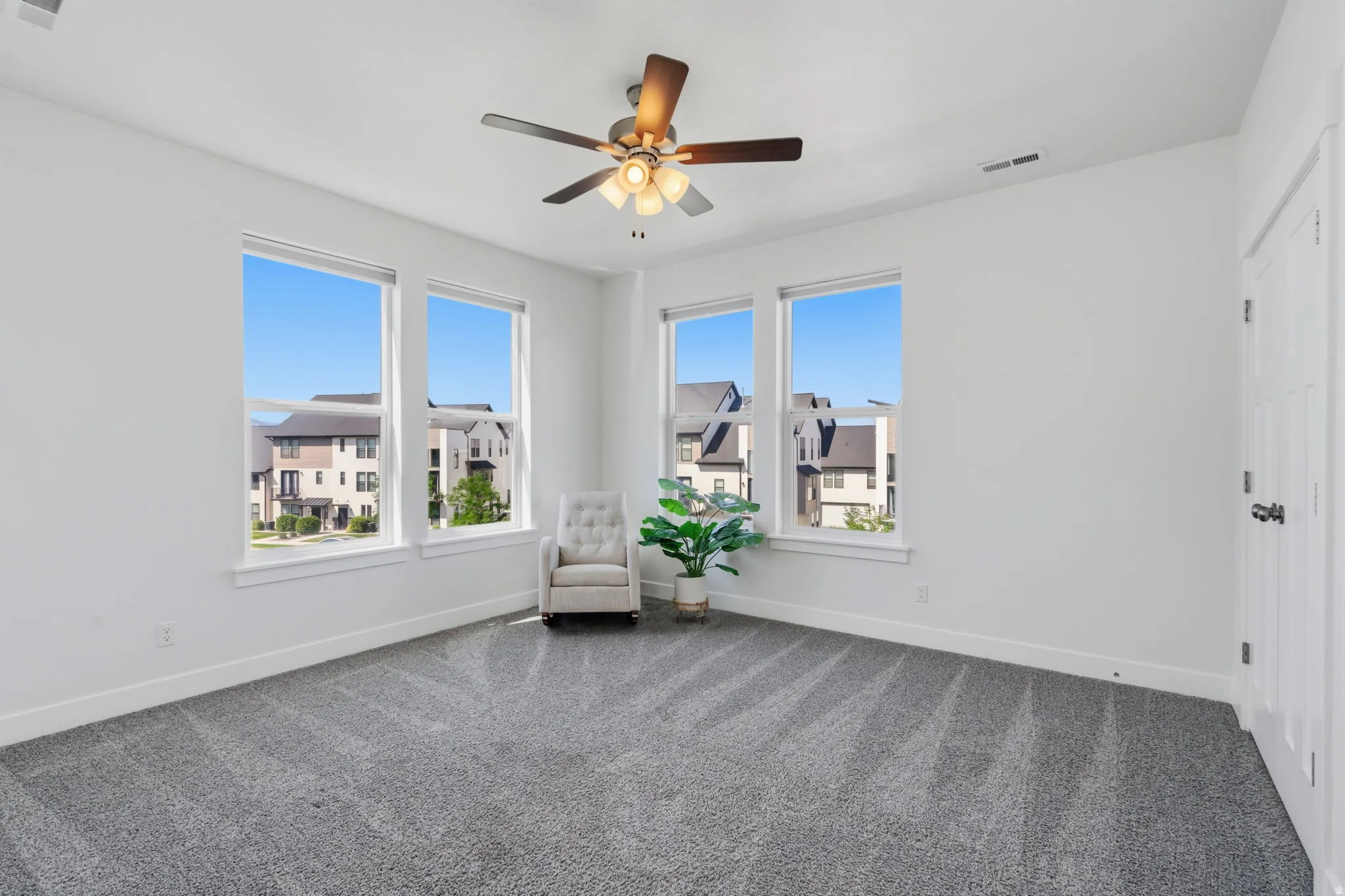 Spacious room featuring multiple large windows, neutral gray carpet, white walls, and a ceiling fan with integrated lighting