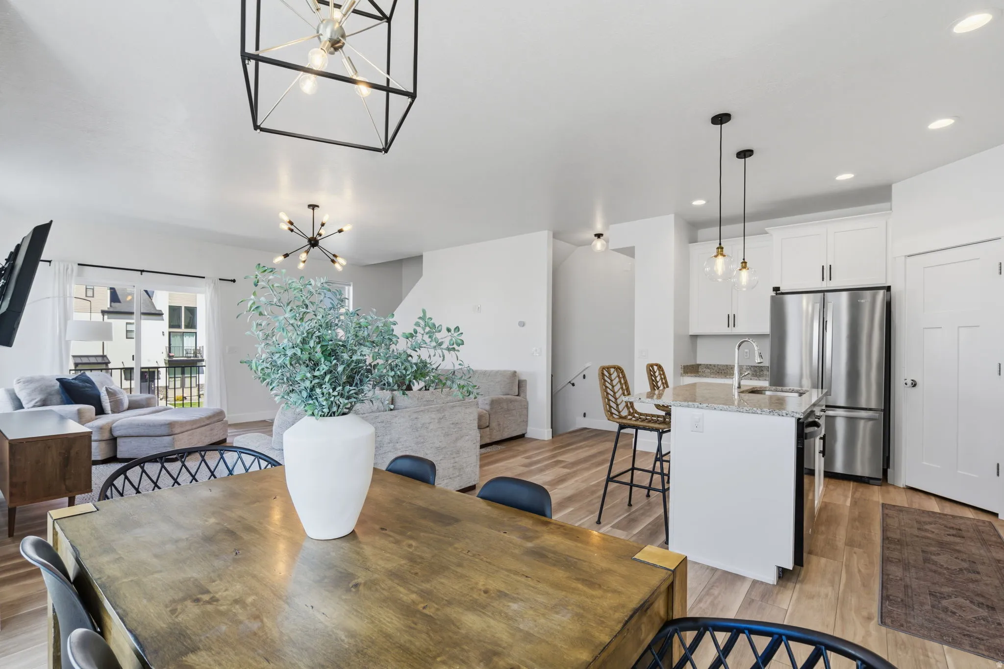 Open-concept living space featuring wood-finish flooring, a contemporary kitchen island with pendant lighting, white shaker cabinetry, stainless steel appliances, and a sliding glass door