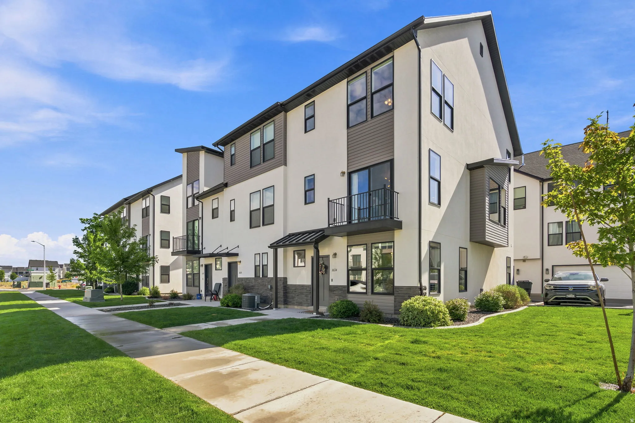 Contemporary townhouse exterior featuring a white stucco finish, dark gray siding, multiple large windows, a private balcony with a metal railing, and an attached garage